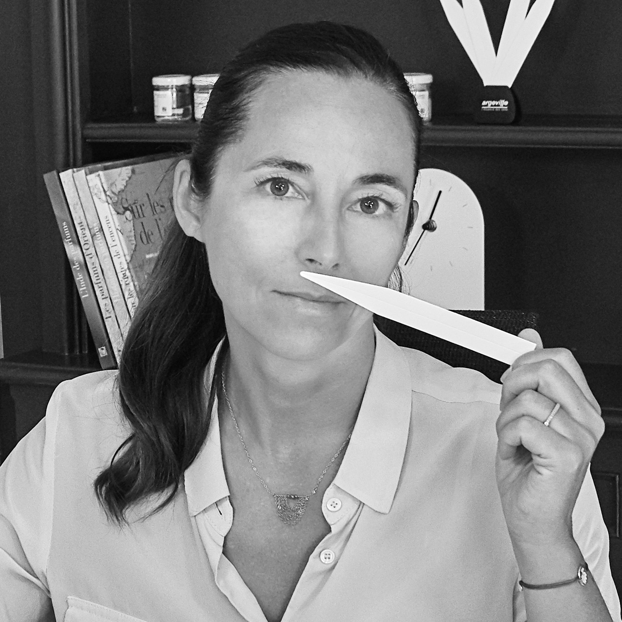 A woman with dark hair in a ponytail holds a paper scent strip near her nose, seated at a desk with books and small jars in the background. The photo is in black and white.
